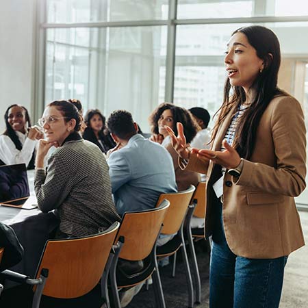 Standing white female presenting to a group seated around a table